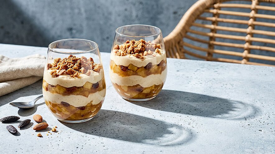 Zwei Gläser mit geschichtetem Dessert sind auf einem Tisch platziert. Die Schichten bestehen aus cremigem Pudding, Äpfeln und Rosinen, gekrönt mit einem knusprigen Kekscrumb. Neben den Gläsern liegen einige Mandeln und ein Löffel auf einer Tischdecke.