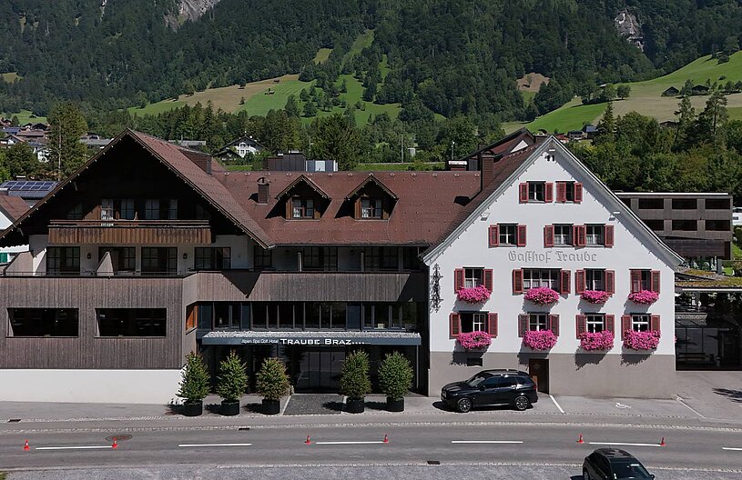 Das Bild zeigt den Gasthof Traube, ein Gebäude mit zwei unterschiedlichen Fassaden. Die linke Seite ist aus Holz, während die rechte Wand weiß ist und mit roten Fensterläden sowie Blumenkästen geschmückt ist. Im Vordergrund sind Bäume und ein Parkplatz zu sehen.