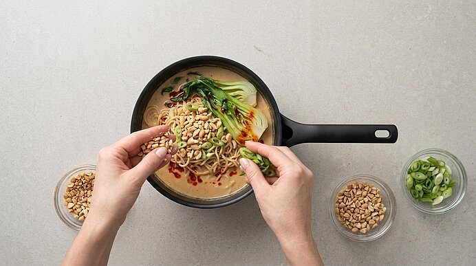 Zwei Hände bestreuen Ramen und Pak Choi in einer Pfanne mit gehackten Erdnüssen und Frühlingszwiebeln.
