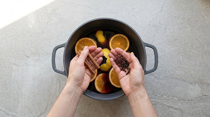 Zwei Hände geben Zimtstangen und Sternanis in einen Topf mit Orangenhälften und Zitrusschalen.