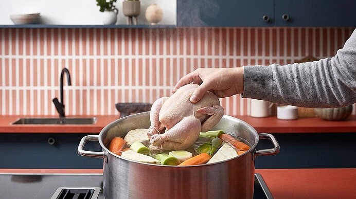 Eine Hand legt ein ganzes rohes Hähnchen in einen großen Topf mit Suppengemüse (Karotten, Lauch, Sellerie) auf dem Herd.