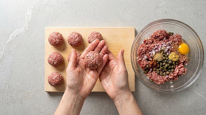 Zwei Hände formen aus gewürztem Hackfleisch ein Fleischbällchen; mehrere fertige Bällchen liegen auf einem Holzbrett, daneben eine Glasschüssel mit rohem Hackfleisch, Ei, Kapern und Gewürzen.