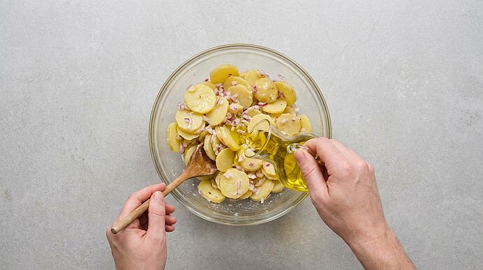Hände: Eine gießt Olivenöl in eine Glasschüssel mit in Scheiben geschnittenen Kartoffeln und roten Zwiebelringen, die andere rührt mit einem Holzlöffel.