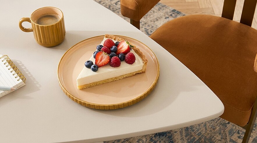 Scheibe Käsekuchen auf einem Teller, garniert mit Erdbeeren, Himbeeren und Heidelbeeren; daneben eine Tasse Kaffee.