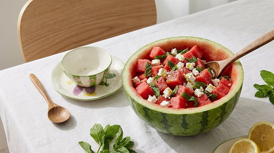 Halbierte Wassermelone als Schüssel mit Melonenwürfeln, zerbröseltem Feta und frischer Minze, angerichtet als Sommersalat auf weißem Tuch; ein Holzlöffel steckt in der Melone, daneben Teetasse und Zitronenscheiben.
