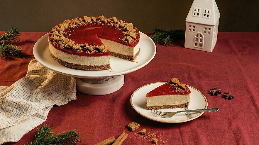 Ein runder Käsekuchen auf einem Teller, bedeckt mit roter Gelee-Oberschicht und Keksstückchen. Ein Stück des Kuchens ist auf einem separaten Teller angerichtet, daneben ein Löffel. Im Hintergrund liegt eine helle Serviette und ein kleines Haus aus Keramik.