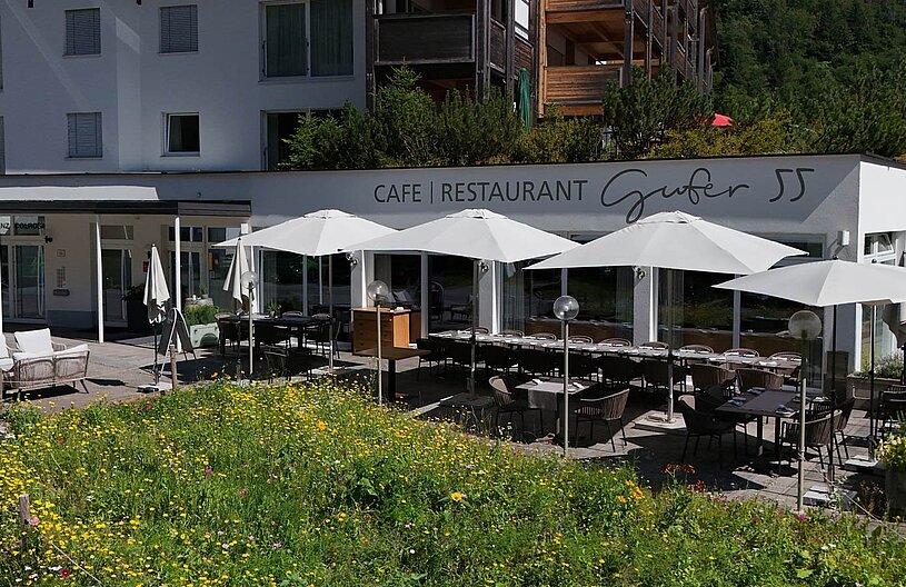 Das Bild zeigt das Café Restaurant Gufer 55 mit einer einladenden Terrasse, die mit Sonnenschirmen und Tischen ausgestattet ist. Vor dem Gebäude wächst eine bunte Blumenwiese. Die Fassade des Restaurants ist modern und freundlich gestaltet.