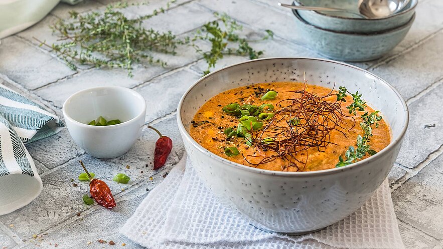 Eine große Schüssel mit cremiger Suppe, garniert mit Frühlingszwiebeln und Kräutern. Neben der Schüssel steht eine kleine Schale mit gehackten Frühlingszwiebeln. Rote Chilischoten und Gewürze sind ebenfalls rund um die Schüssel verteilt, auf einem hellen Untergrund.