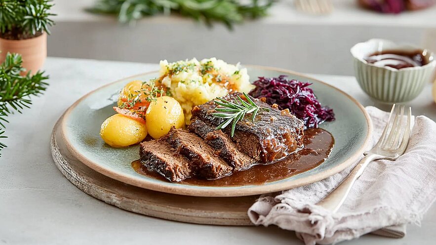 Auf dem Teller befindet sich ein saftiges Stück Rindfleisch, garniert mit frischen Kräutern. Dazu gibt es goldene Kartoffeln, cremigen Kartoffelbrei und Rotkohl. Die Speisen sind appetitlich angerichtet und umgeben von einem rustikalen Teller.