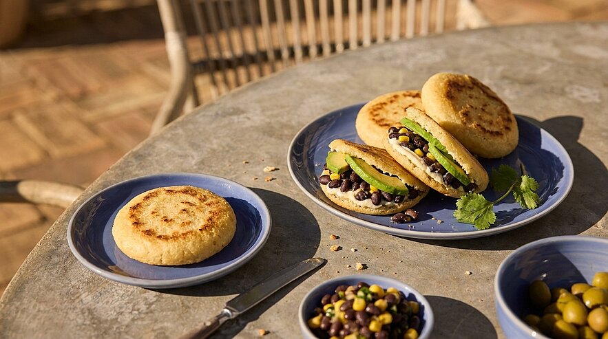 Auf einem Tisch liegen mehrere gefüllte Arepas auf blauen Tellern, darunter schwarze Bohnen und frisches Gemüse. Zusätzlich befindet sich eine einzelne Arepa auf einem kleineren Teller. Der Tisch hat eine rustikale Oberfläche und die Atmosphäre wirkt einladend.