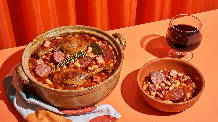 Topf mit Cassoulet: weißer Bohnen-Eintopf mit Entenkeulen, Wurst- und Speckstücken, garniert mit Thymian; daneben eine Schale mit Portion und ein Glas Rotwein.