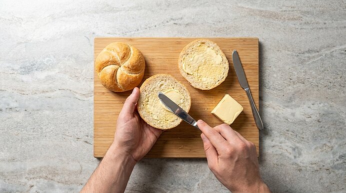 Zwei Hände bestreichen die untere Hälfte einer aufgeschnittenen Semmel auf einem Holzbrett mit Butter.