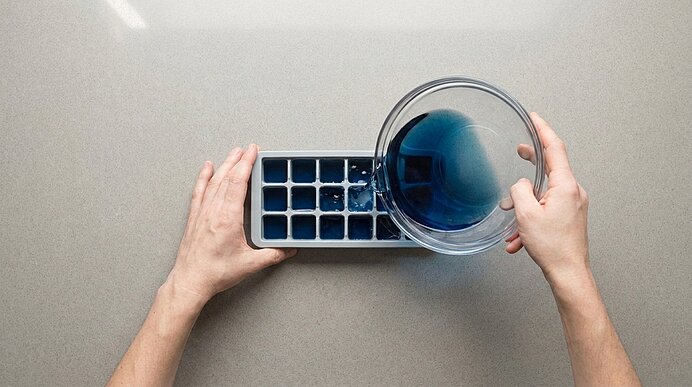 Zwei Hände gießen aus einer Glasschale blaue Flüssigkeit in eine Eiswürfelform auf einer hellen Arbeitsfläche.