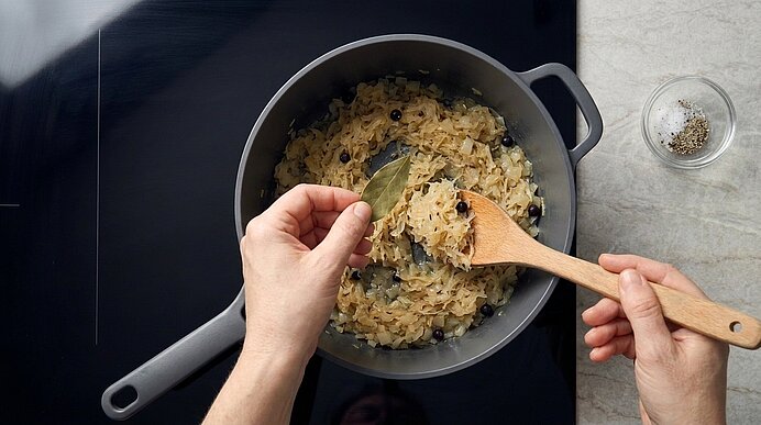 Hände legen ein Lorbeerblatt in einen Topf mit gedünstetem Kraut und rühren mit einem Holzlöffel.