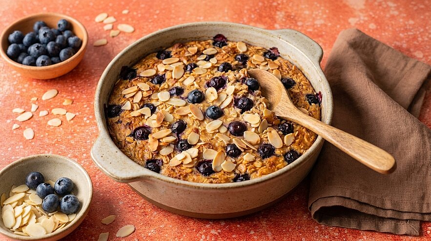 Keramikschale mit gebackenem Haferauflauf, bestreut mit Blaubeeren und Mandelblättchen; ein Holzlöffel liegt in der Schale.
