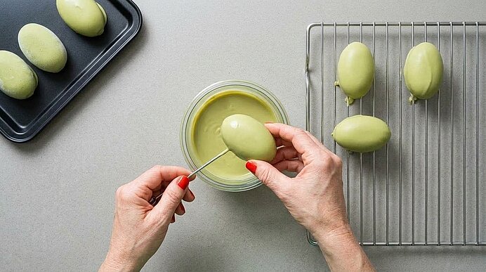 Zwei Hände tauchen ein gefrorenes Pistaziendessert an einer Nadel in eine Schüssel mit hellgrüner Glasur.