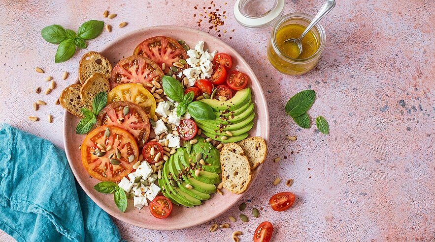 Teller mit sommerlichem Salat: unterschiedlich große Tomatenscheiben, Avocadoscheiben, halbierte Kirschtomaten, zerbröseltem Feta, frischen Basilikumblättern und gerösteten Brotscheiben; kleines Glas Öl‑Dressing.