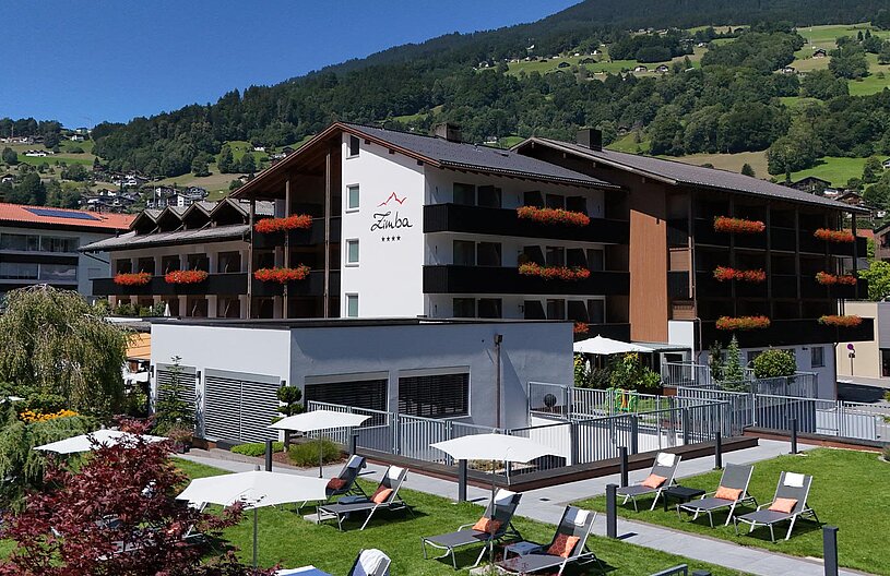 Das Hotel Zimba liegt in einer malerischen Umgebung, umgeben von Bergen und Grünflächen. Die Fassade des Hotels ist modern mit Blumenkästen. Im Vordergrund befinden sich Liegen auf einem gepflegten Rasen und Sonnenschirme, die eine einladende Atmosphäre schaffen.
