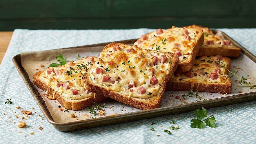 Ein Blech mit mehreren Scheiben Toastbrot, überbacken mit Käse, Schinken und Kräutern. Die knusprigen Toasts sind goldbraun und werden auf Pergamentpapier präsentiert, umgeben von einigen frischen Kräutern.