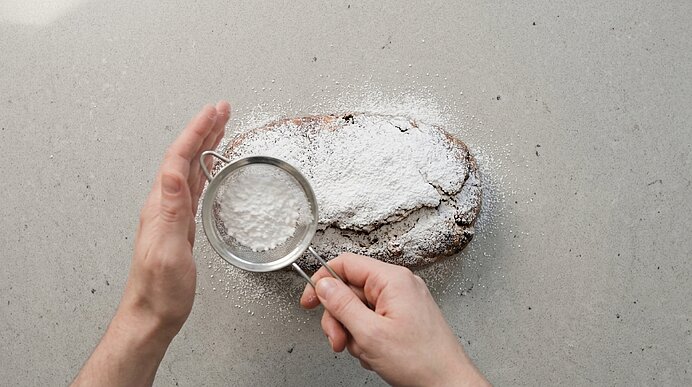 Zwei Hände streuen Puderzucker mit einem feinen Sieb über einen länglichen Kuchenlaib auf hellem Untergrund.