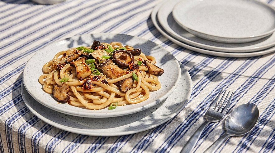 Teller mit gebratenen Udon-Nudeln, gebratenen Pilzen (Shiitake), knusprigen Tofuwürfeln, Frühlingszwiebeln, Sesam und scharfer Chili-Öl-Sauce.