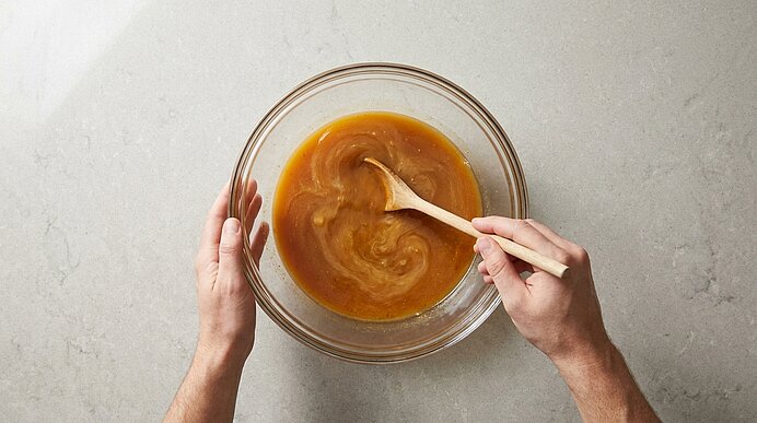 Zwei Hände rühren mit einem Holzlöffel eine goldbraune Karamellsauce in einer großen Glasschüssel auf hellem Untergrund.