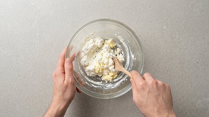 Zwei Hände verrühren Topfen und Butter mit einem Holzlöffel in einer Glasschüssel auf hellgrauer Arbeitsfläche.