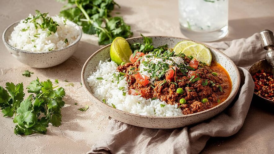 Eine Schüssel mit Reis und einer würzigen Fleischsoße, die mit frischen Kräutern, Tomatenstücken und einem Klecks Joghurt garniert ist. Lime-Scheiben liegen auf dem Gericht. Im Hintergrund sind zusätzliche Schalen mit Reis und frischen Kräutern zu sehen.