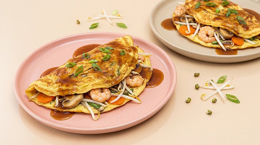 Zwei herzhafte Omeletts auf Tellern, jeweils gefüllt mit Garnelen, Mungobohnensprossen, Pilzen und Karotten, garniert mit dunkler Sauce und Frühlingszwiebeln.
