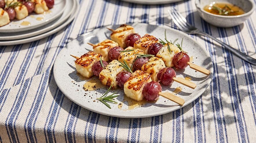 Ein Teller mit gegrillten Spießen aus Käse und Trauben, garniert mit frischem Rosmarin. Die Spieße liegen auf einem gestreiften Tischcloth. Im Hintergrund sind weitere Teller und eine kleine Schale mit Sauce zu sehen.