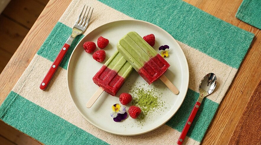 Zwei zweifarbige Eis am Stiel (grün oben, rot unten) auf einem weißen Teller, garniert mit frischen Himbeeren, etwas grünem Pulver (Matcha) und kleinen essbaren Blüten.