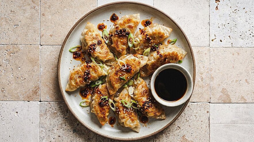 Teller mit goldbraun gebratenen Teigtaschen (Gyoza), garniert mit Frühlingszwiebeln, Sesam und Chiliöl, daneben eine kleine Schale dunkler Sojasauce zum Dippen.