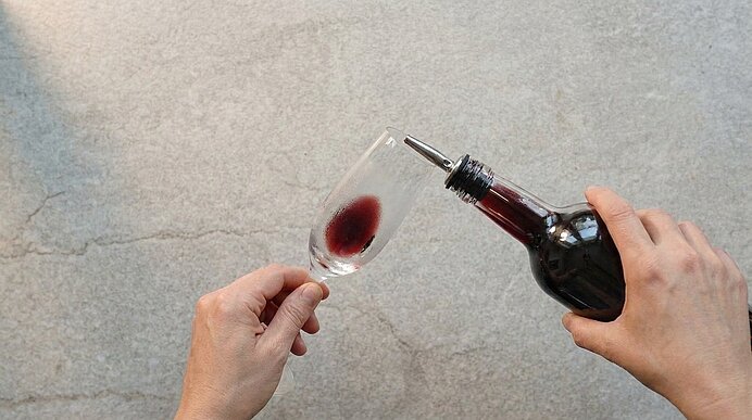 Zwei Hände gießen dunklen Crème de Cassis aus einer Flasche in ein gekühltes Sektglas.