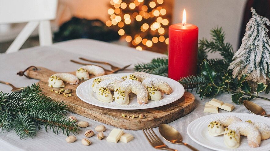 Teller mit Vanillekipferl, an den Enden in weiße Schokolade getaucht und mit gehackten Pistazien bestreut, auf einem weihnachtlichen Tisch mit brennender roter Kerze und Tannenzweigen.