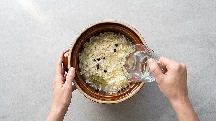 Zwei Hände gießen Wasser in eine Schüssel mit fein geschnittenem Weißkohl, Lorbeerblättern und Pfefferkörnern zum Einlegen.