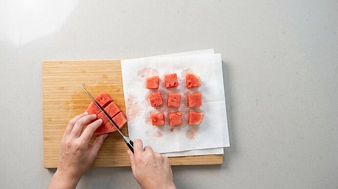Zwei Hände schneiden Wassermelone in Würfel. Daneben tropfen geschnittene Würfel auf Küchenpapier ab.