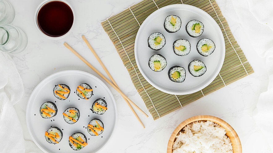 Zwei weiße Teller mit Sushi-Rollen auf einem Bambusmatte. Eine Platte enthält Sushi mit Gemüse und eine andere mit Avocado. Daneben steht eine Schale mit gekochtem Reis und ein Getränk in einem Glas. Essstäbchen liegen bereit.