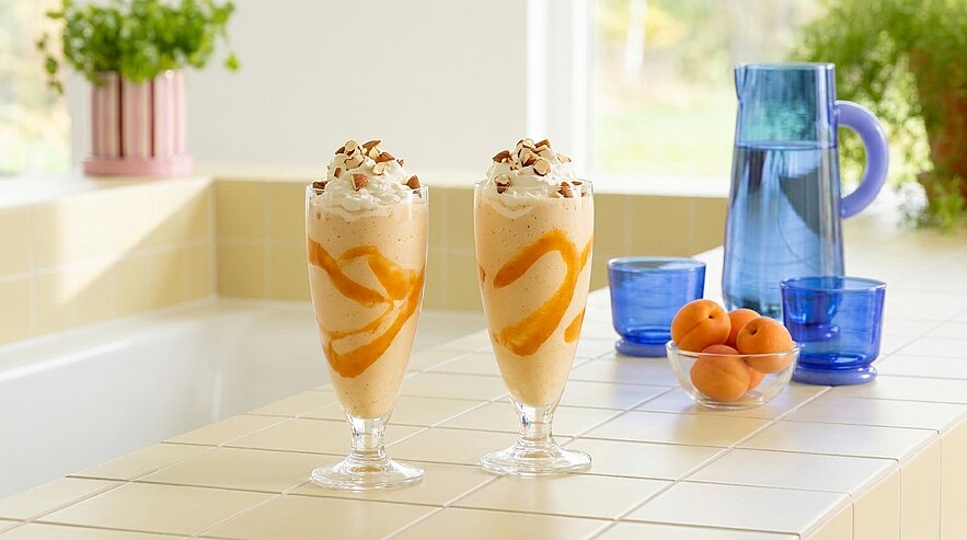 Zwei hohe Dessertgläser mit aprikosenfarbener Eismousse und orangenfarbener Sauce, garniert mit Schlagrahm und gehackten Nüssen, stehen auf einer hellen Küchenarbeitsplatte.