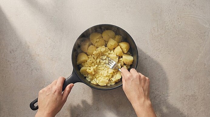 Zwei Hände zerdrücken in einem schwarzen Topf mit einer Gabel gekochte Kartoffelstücke zu Kartoffelstampf.