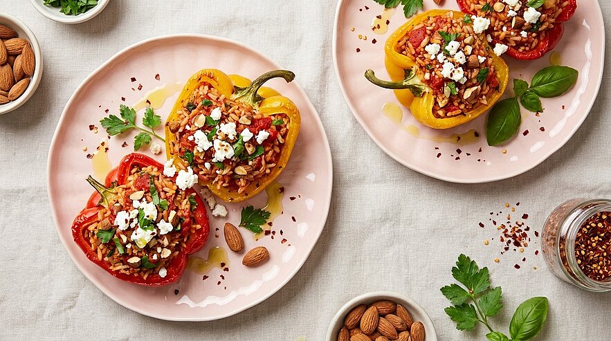 Zwei Teller mit gefüllten Paprikaschoten (rot und gelb), gefüllt mit Reis, Tomaten, Kräutern und zerbröseltem Feta, garniert mit Petersilie.
