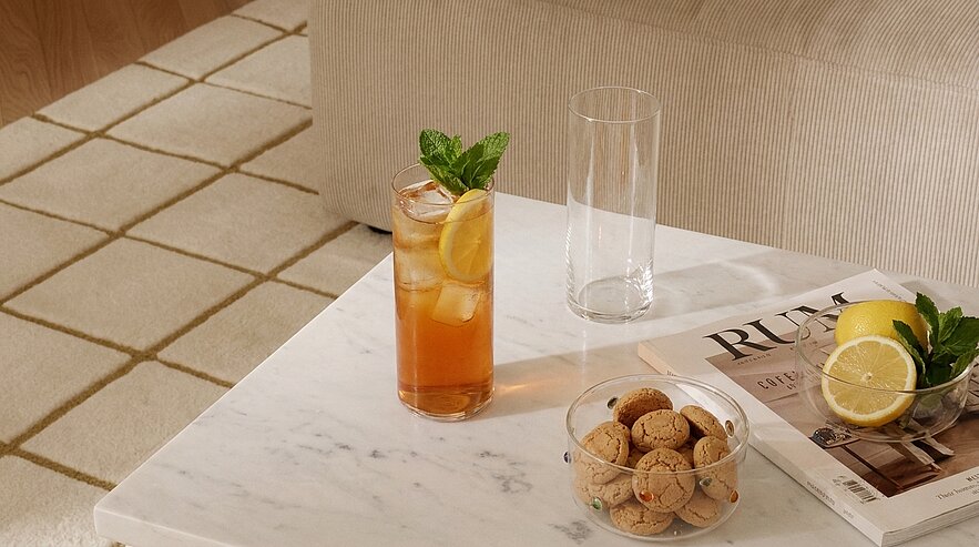 Weißer Marmortisch mit einem Glas Eistee mit Zitronenscheibe und Minze, daneben ein leeres Glas, eine Schale mit kleinen Keksen und eine halbierte Zitrone auf einem Magazin.