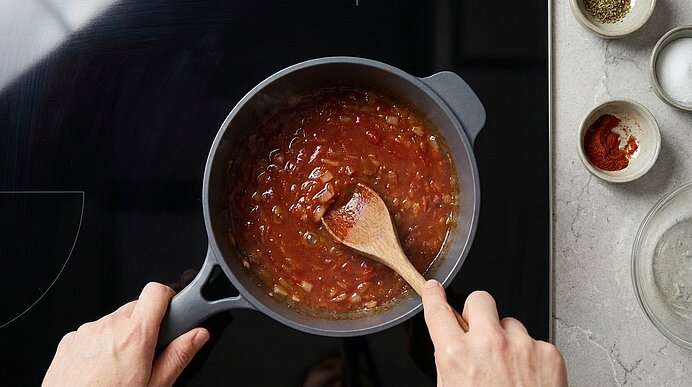 Hände rühren mit einem Holzlöffel eine Tomatensoße in einem Topf auf dem Herd; rechts stehen kleine Schalen mit Gewürzen.