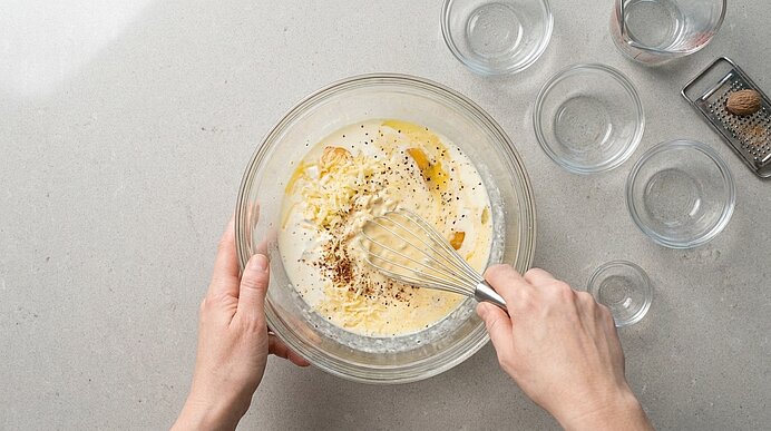 Zwei Hände verquirlen Eier, Sahne, Käse und Gewürze in einer Glasschüssel auf hellgrauer Stein-Arbeitsplatte.