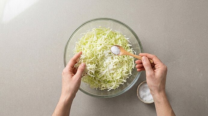 Zwei Hände streuen mit einem kleinen Holzlöffel Salz über eine Glasschüssel mit frisch gehobeltem Weißkohl.