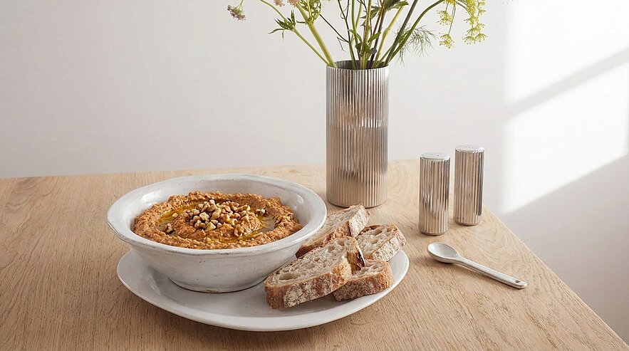 Eine Schüssel mit Hummus, garniert mit Nüssen, steht auf einem weißen Teller auf einem Holztisch. Daneben liegen ein paar Stücke Brot und ein kleiner Löffel. Im Hintergrund ist eine Vase mit frischen Blumen zu sehen.