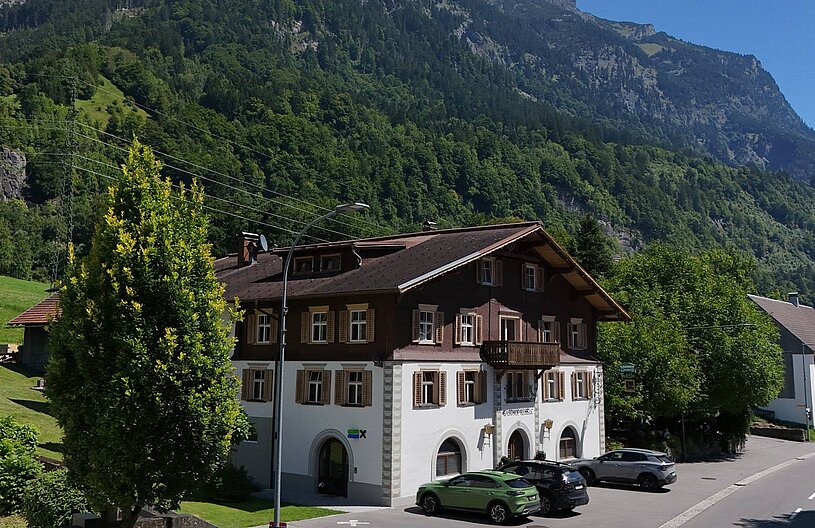 Ein traditionelles Haus mit Holzfassade steht in einer grünen Landschaft, umgeben von Bäumen und Bergen. Auf der Straße parken mehrere Autos. Die Sonne scheint und der Himmel ist klar.