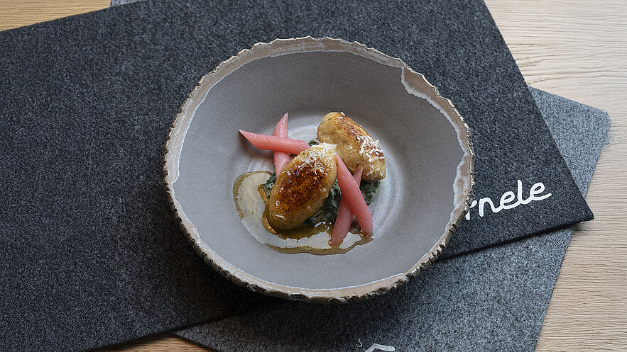 Ein tiefes, handgefertigtes Geschirr enthält zwei goldbraun gebratene Kugeln aus Kartoffelteig, die auf einem Bett aus Blattspinat liegen. Dazwischen sind pinkfarbene Rhabarberstreifen angerichtet, mit einer feinen Sauce umgeben. Der Hintergrund besteht aus grauen Tischunterlagen.