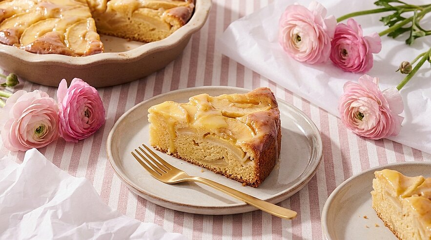 Ein Stück Apfelkuchen auf einem Teller mit einer goldenen Gabel. Im Hintergrund steht eine Torte in einer runden Form. Blüten in sanften Farben und leicht zerknittertes Papier ergänzen das Arrangement.