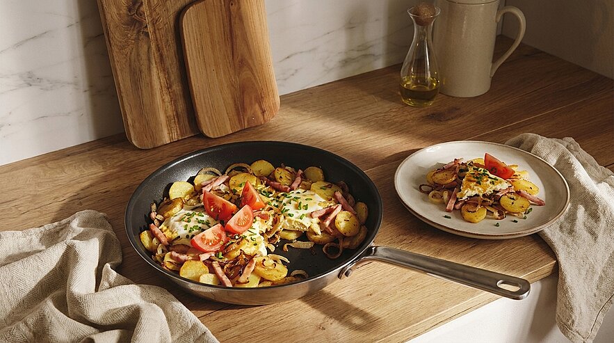 Pfanne mit goldbraunen Bratkartoffeln, gebratenem Speck, Spiegelei, Tomatenvierteln und Schnittlauch; daneben ein Teller mit einer Portion.
