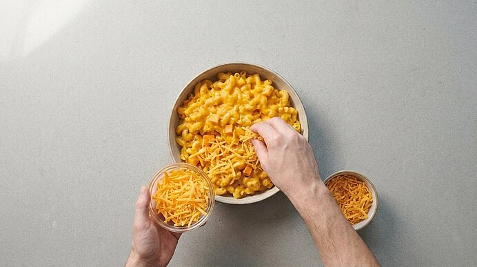 Zwei Hände bestreuen Kürbis-Mac-and-Cheese in einer Form mit geriebenem Cheddar.